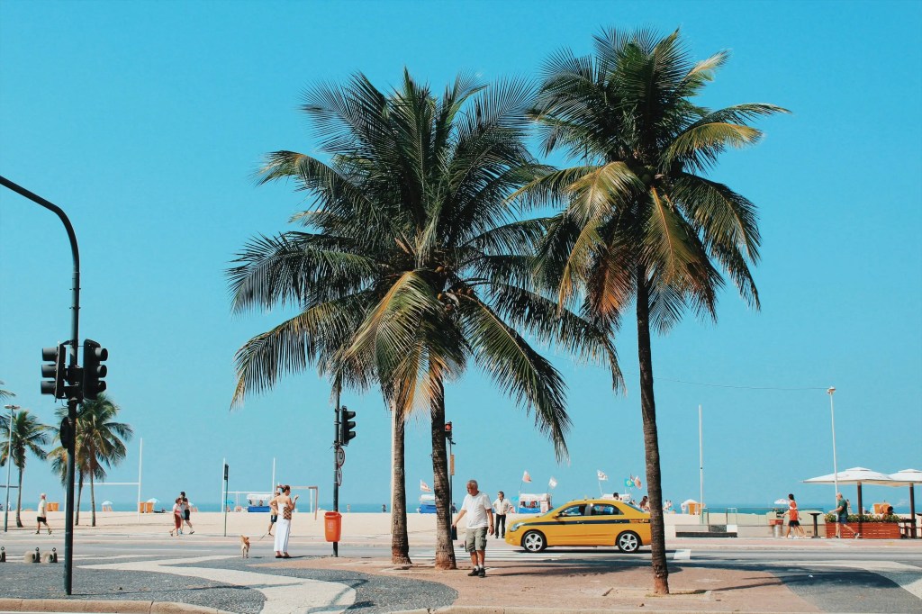 Taxi amarelo na orla de Copacabana com palmeiras e calçadão, representando a Zona Sul do Rio.