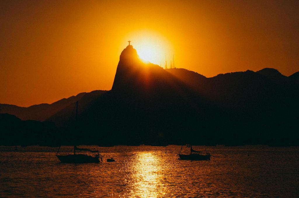 Silhueta do Cristo Redentor no pôr do sol, com o céu alaranjado e barcos na Baía de Guanabara.